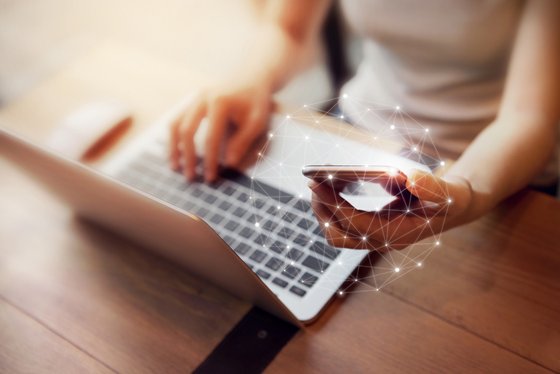 An open laptop with hands, one holding a phone out with a network graphic around it.